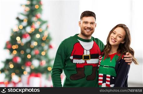 winter holidays, celebration and people concept - portrait of happy couple in ugly sweaters at home over christmas tree lights background. happy couple in ugly sweaters over christmas tree