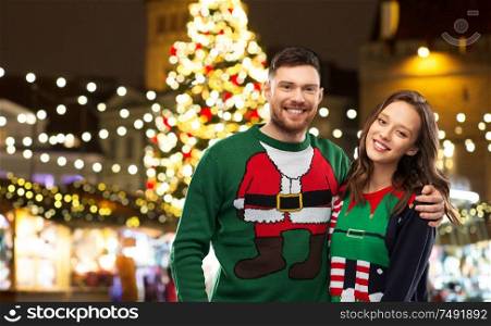 winter holidays, celebration and people concept - portrait of happy couple in ugly sweaters hugging over christmas market lights background. happy couple in ugly sweaters at christmas market