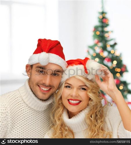 winter, holidays, celebration and people concept - smiling couple in sweaters and santa helper hats over living room with christmas tree background