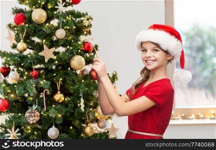 winter holidays, celebration and people concept - smiling girl in santa helper hat decorating christmas tree at home. happy girl in santa hat decorating christmas tree