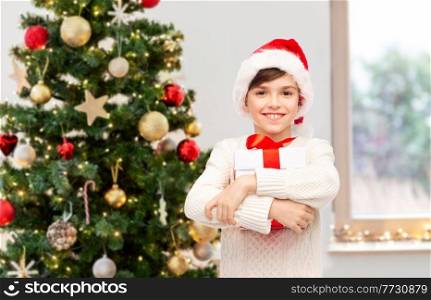 winter holidays, childhood and people concept - smiling happy boy in santa hat with gift box over christmas tree background. smiling happy boy in santa hat with gift box
