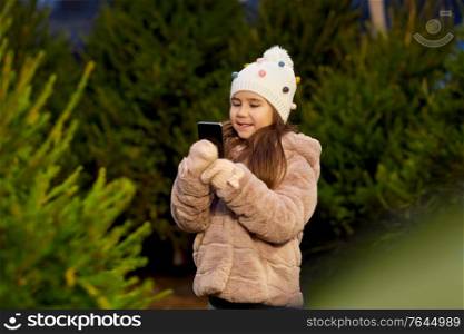 winter holidays, children and people concept -happy smiling little girl with smartphone at christmas tree market in evening. girl with smartphone at christmas tree market
