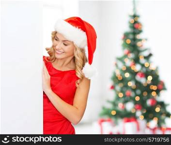 winter holidays, christmas, advertising and people concept - smiling young woman in santa helper hat with white blank billboard over living room with christmas tree background