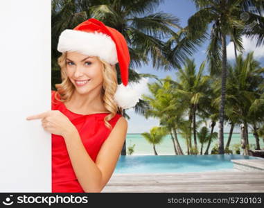 winter holidays, christmas, advertising and people concept - smiling young woman in santa helper hat with white blank billboard over tropical beach and swimming pool background