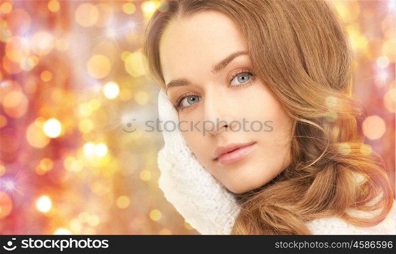 winter, holidays, christmas and people concept - close up of happy young woman in mittens touching her face over lights background