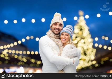 winter, holidays, christmas and people concept - happy couple in hats and scarf hugging over night lights background. happy couple hugging over christmas lights