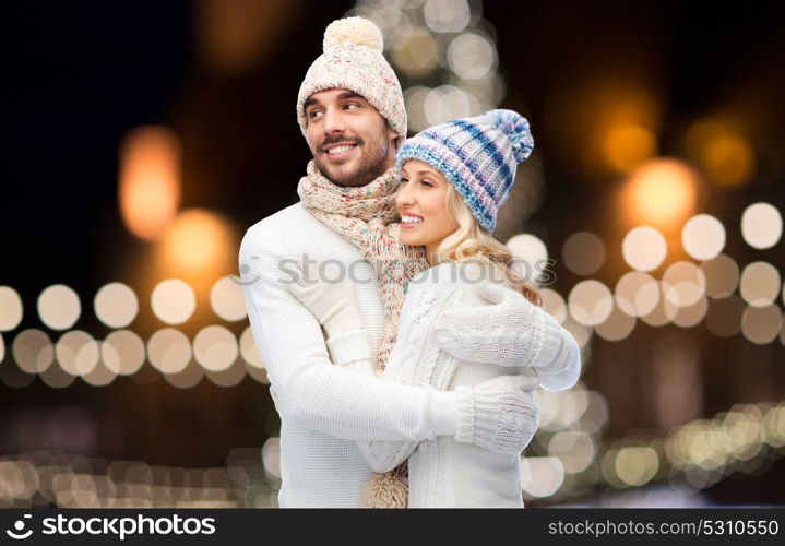 winter, holidays, christmas and people concept - happy couple in hats and scarf hugging over night lights background. happy couple hugging over christmas lights