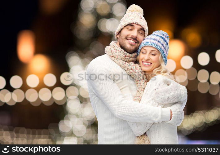 winter, holidays, christmas and people concept - happy couple in hats and scarf hugging over night lights background. happy couple hugging over christmas lights