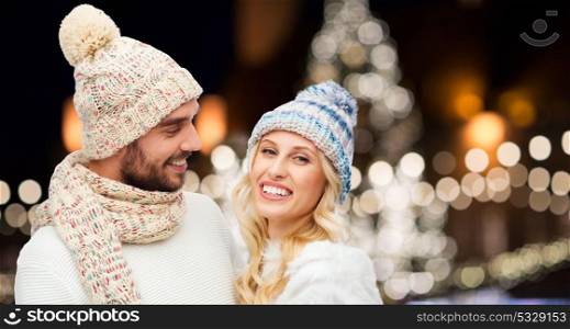 winter, holidays, christmas and people concept - happy couple in hats and scarf hugging over night lights background. happy couple hugging over christmas lights