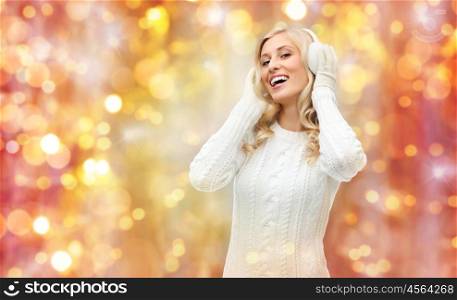 winter, holidays, christmas and people concept - smiling young woman in earmuffs and sweater over lights background