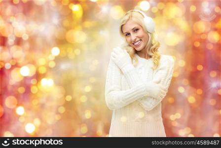 winter, holidays, christmas and people concept - smiling young woman in earmuffs and sweater over lights background