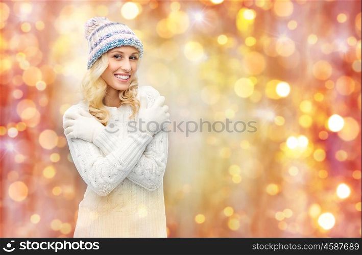 winter, holidays, christmas and people concept - smiling young woman in winter hat, sweater and gloves over lights background