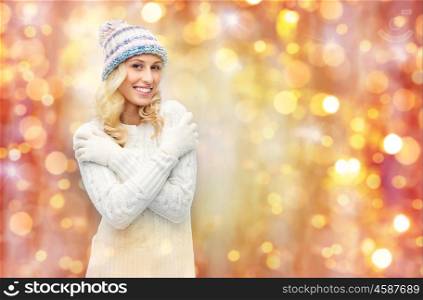 winter, holidays, christmas and people concept - smiling young woman in winter hat, sweater and gloves over lights background