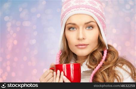 winter holidays, christmas, beverages and people concept - happy young woman in winter hat with red cup of tea over rose quartz and serenity lights background