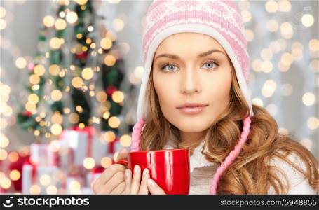 winter holidays, christmas, beverages and people concept - happy young woman in winter hat with red cup over christmas tree lights background
