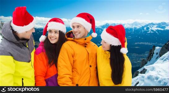 winter holidays, christmas, friendship and people concept - happy friends in santa hats and ski suits talking outdoors over snowy mountains background