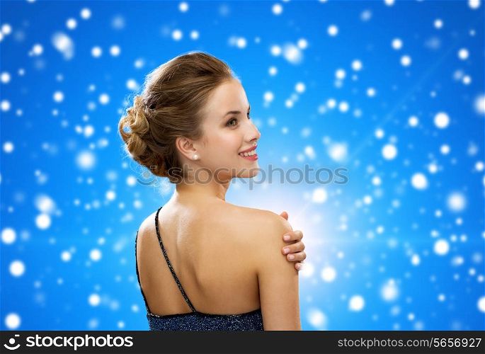winter holidays, christmas, people and glamour concept - smiling woman in evening dress over black background over blue snowy background from back