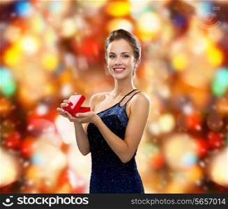 winter holidays, christmas, presents, luxury and happiness concept - smiling woman in dress holding red gift box over red lights background
