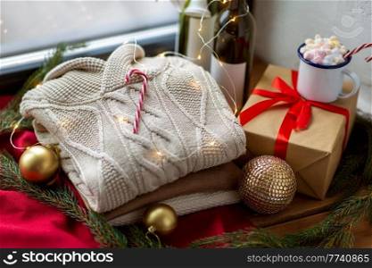 winter holidays concept - close up of warm wool braided sweater, candy cane, electric garland string, christmas decorations and candle burning in wine bottle on window sill at home. close up of sweater and christmas decor on sill