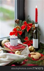 winter holidays concept - cup of whipped cream with marshmallow and candy canes, books, oatmeal cookies, candle burning in wine bottle and christmas decorations on window sill at home. christmas treats and decorations on window sill