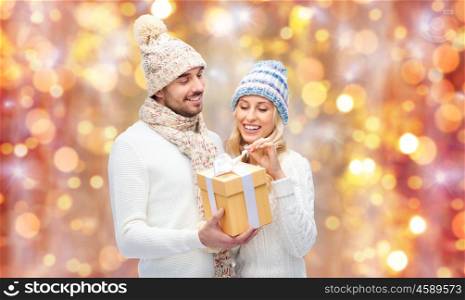winter, holidays, couple, christmas and people concept - smiling man and woman in hats and scarf with gift box over lights background