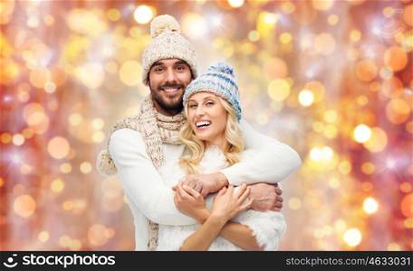winter, holidays, couple, christmas and people concept - smiling man and woman in hats and scarf hugging over lights background