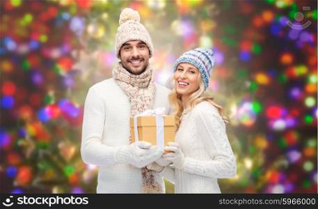 winter, holidays, couple, christmas and people concept - smiling man and woman in hats and scarf with gift box over holidays lights background