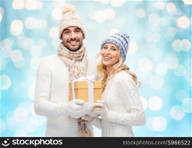 winter, holidays, couple, christmas and people concept - smiling man and woman in hats and scarf with gift box over blue holidays lights background