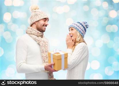 winter, holidays, couple, christmas and people concept - smiling man and woman in hats and scarf with gift box over blue holidays lights background