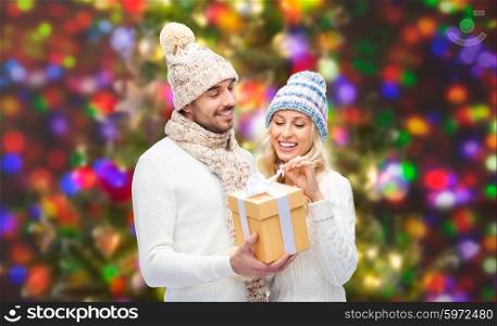 winter, holidays, couple, christmas and people concept - smiling man and woman in hats and scarf with gift box over lights background