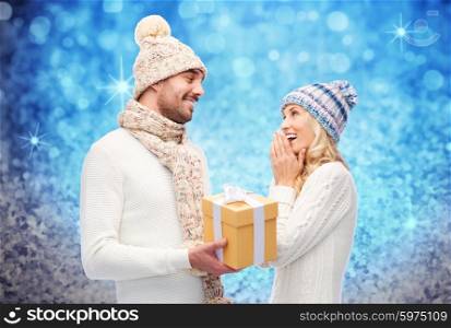 winter, holidays, couple, christmas and people concept - smiling man and woman in hats and scarf with gift box over blue glitter or lights background