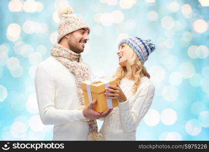 winter, holidays, couple, christmas and people concept - smiling man and woman in hats and scarf with gift box over blue holidays lights background