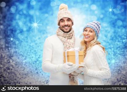 winter, holidays, couple, christmas and people concept - smiling man and woman in hats and scarf with gift box over blue glitter and holidays lights background