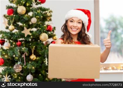 winter holidays, delivery and people concept - smiling woman in santa helper hat with parcel box showing thumbs up over christmas tree on background. happy woman in santa hat with parcel on christmas