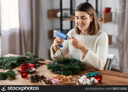 winter holidays, diy and hobby concept - happy smiling woman with hot glue gun and berry decorations making fir christmas wreath at home. woman making fir christmas wreath at home
