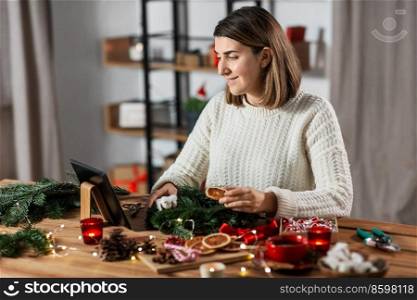 winter holidays, diy and hobby concept - happy smiling woman with tablet pc computer making christmas fir wreath at home. woman with tablet pc makes christmas wreath