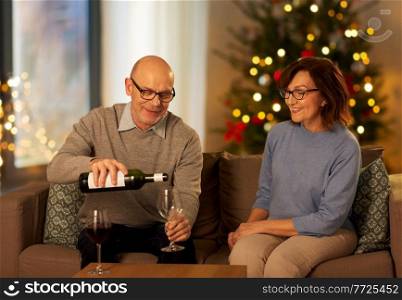 winter holidays, drinks and people concept - happy smiling senior couple drinking red wine at home in evening over christmas tree lights on background. senior couple drinking red wine on christmas
