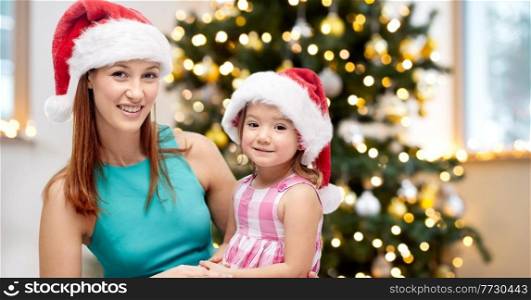 winter holidays, family and people concept - happy mother and little girl in santa hats at home over christmas tree background. happy mother and daughter at home on christmas