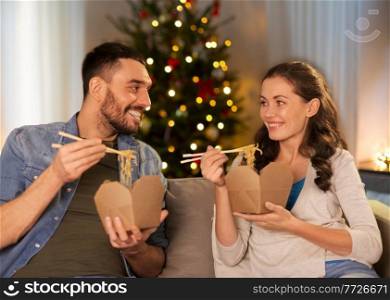 winter holidays, fast food and people concept - happy couple eating takeaway noodles with chopstick at home in evening over christmas tree lights on background. happy couple eating takeaway noodles on christmas
