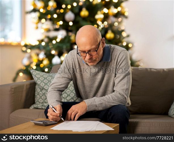 winter holidays, finances and people concept - senior man with papers or bills and calculator writing at home in evening over christmas tree lights on background. senior man with bills at home on christmas
