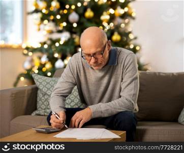 winter holidays, finances and people concept - senior man with papers or bills and calculator writing at home in evening over christmas tree lights on background. senior man with bills at home on christmas