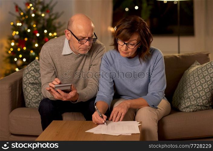 winter holidays, finances and people concept - smiling senior couple with papers and calculator at home over christmas tree lights on background. old couple with bills and calculator on christmas