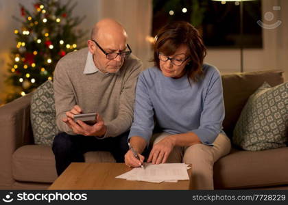 winter holidays, finances and people concept - smiling senior couple with papers and calculator at home over christmas tree lights on background. old couple with bills and calculator on christmas