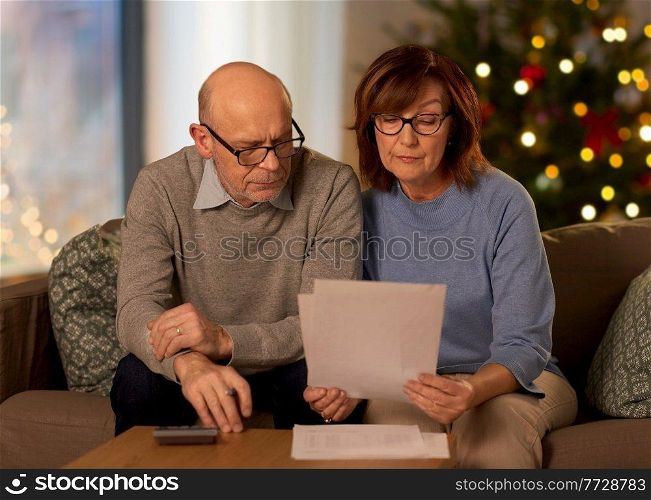 winter holidays, finances and people concept - smiling senior couple with papers and calculator at home over christmas tree lights on background. senior couple with bills at home on christmas