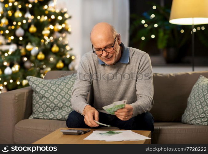 winter holidays, finances and people concept - smiling senior man with calculator and bills counting money at home in evening over christmas tree lights on background. senior man counting money at home on christmas