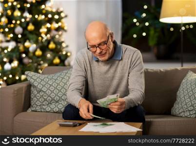 winter holidays, finances and people concept - smiling senior man with calculator and bills counting money at home in evening over christmas tree lights on background. senior man counting money at home on christmas