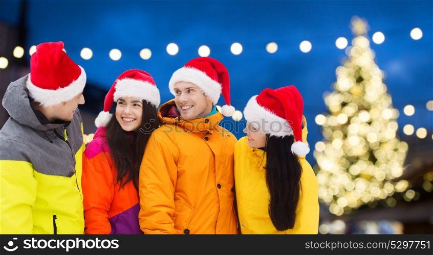 winter holidays, friendship and people concept - happy friends in santa hats and ski suits outdoors over christmas lights background. friends in santa hats and ski suits at christmas