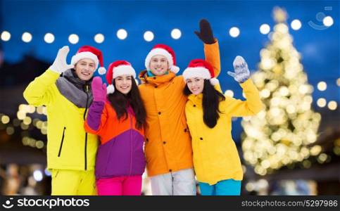 winter holidays, friendship and people concept - happy friends in santa hats and ski suits waving hands outdoors over christmas lights background. friends in santa hats and ski suits at christmas
