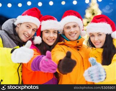 winter holidays, friendship and people concept - happy friends in santa hats and ski suits outdoors showing thumbs up gesture over christmas lights background. happy friends in santa hats and ski suits outdoors