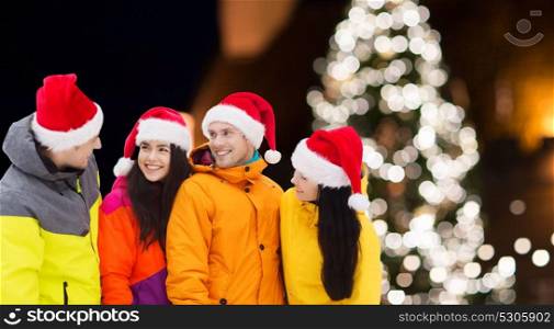 winter holidays, friendship and people concept - happy friends in santa hats and ski suits outdoors over christmas lights background. friends in santa hats and ski suits at christmas